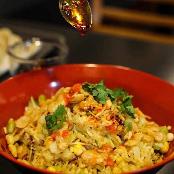 A spoon drizzles chili oil over a vibrant Burmese noodle salad with peanuts and cilantro in a red bowl.