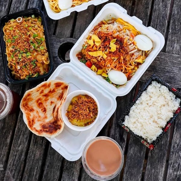 Burmese takeout containers with noodles, rice, paratha, and tea on a wooden table.