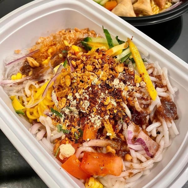 Burmese noodle salad with various toppings, including fried garlic, vegetables, and sauce, in a white container.