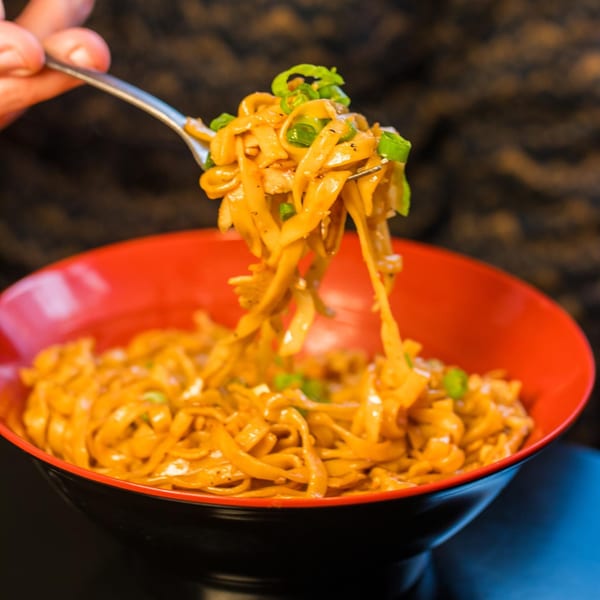A fork lifts saucy Burmese noodles with green onions from a red and black bowl.