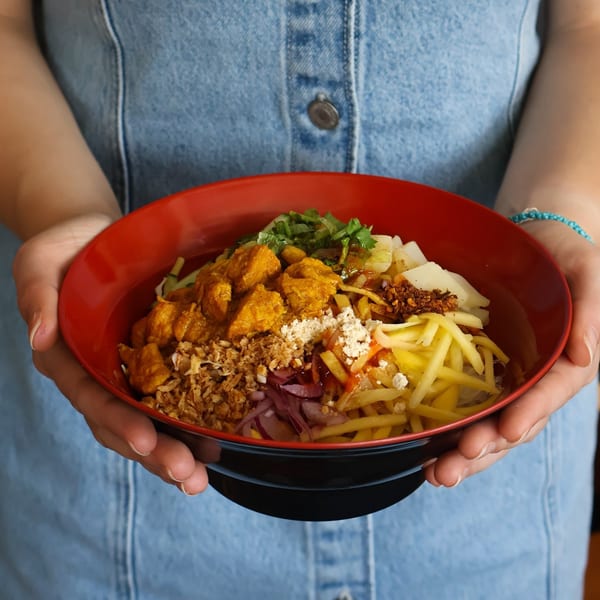 Hands holding a red and black bowl filled with colorful Burmese chicken curry noodles and toppings.
