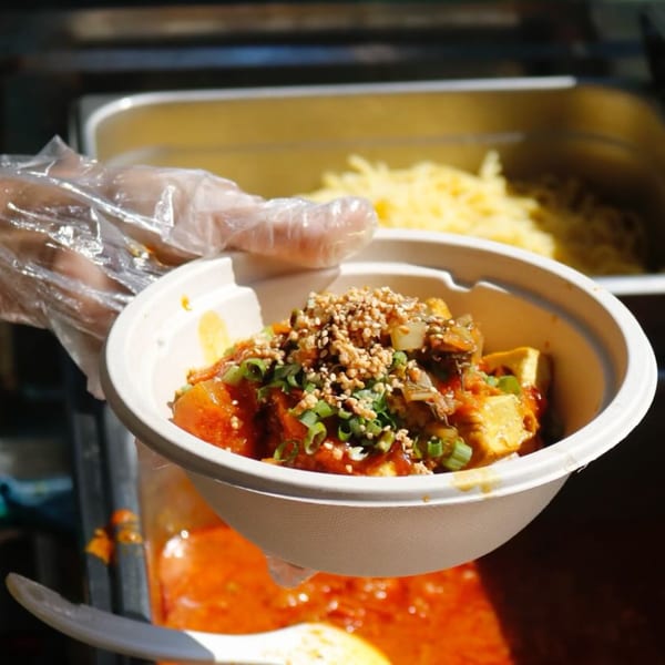 A gloved hand holds a bowl of Burmese tofu with sauce, green onions, and peanuts.