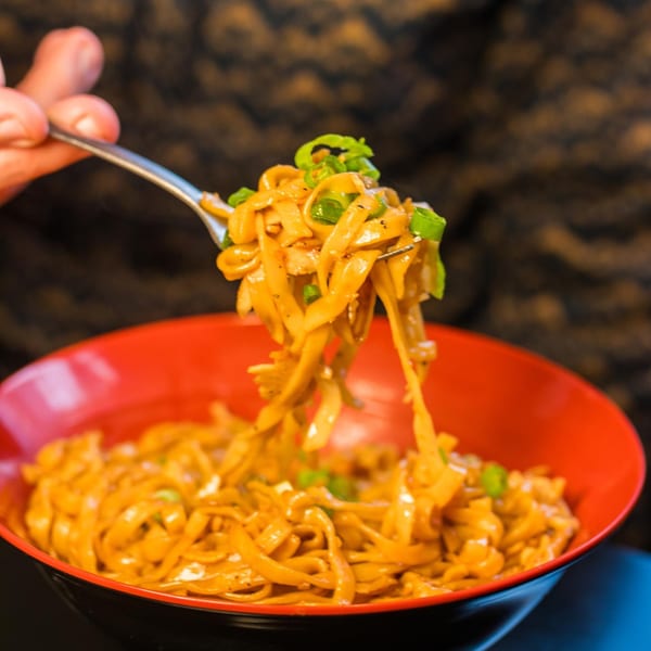 A fork lifts saucy Burmese noodles with green onions from a red and black bowl.