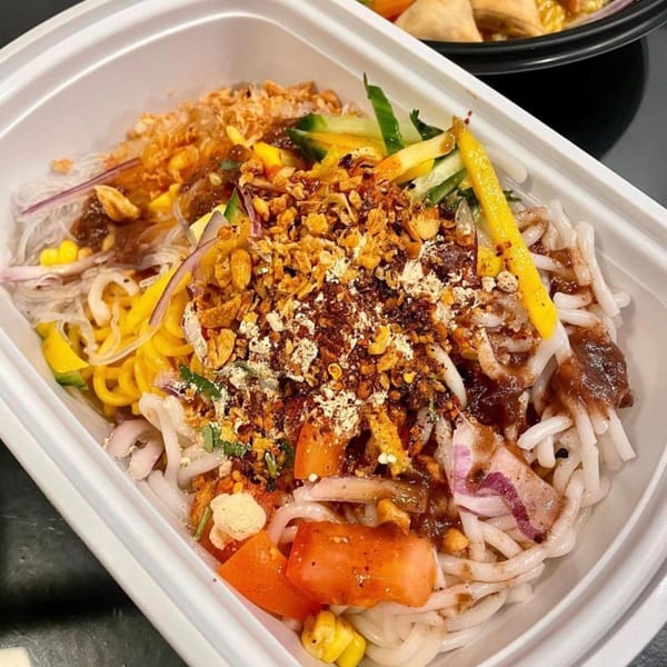 Burmese noodle salad with various toppings, including fried garlic, vegetables, and sauce, in a white container.