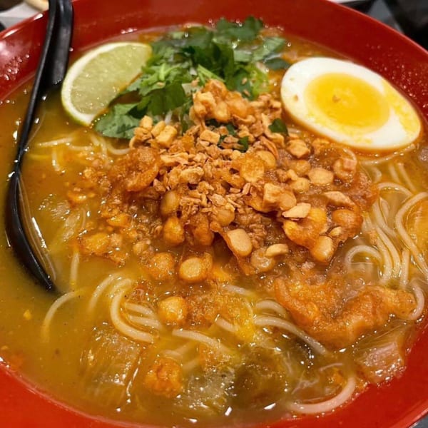 A vibrant red bowl of Burmese mohinga noodle soup with egg, lime, and crispy toppings.